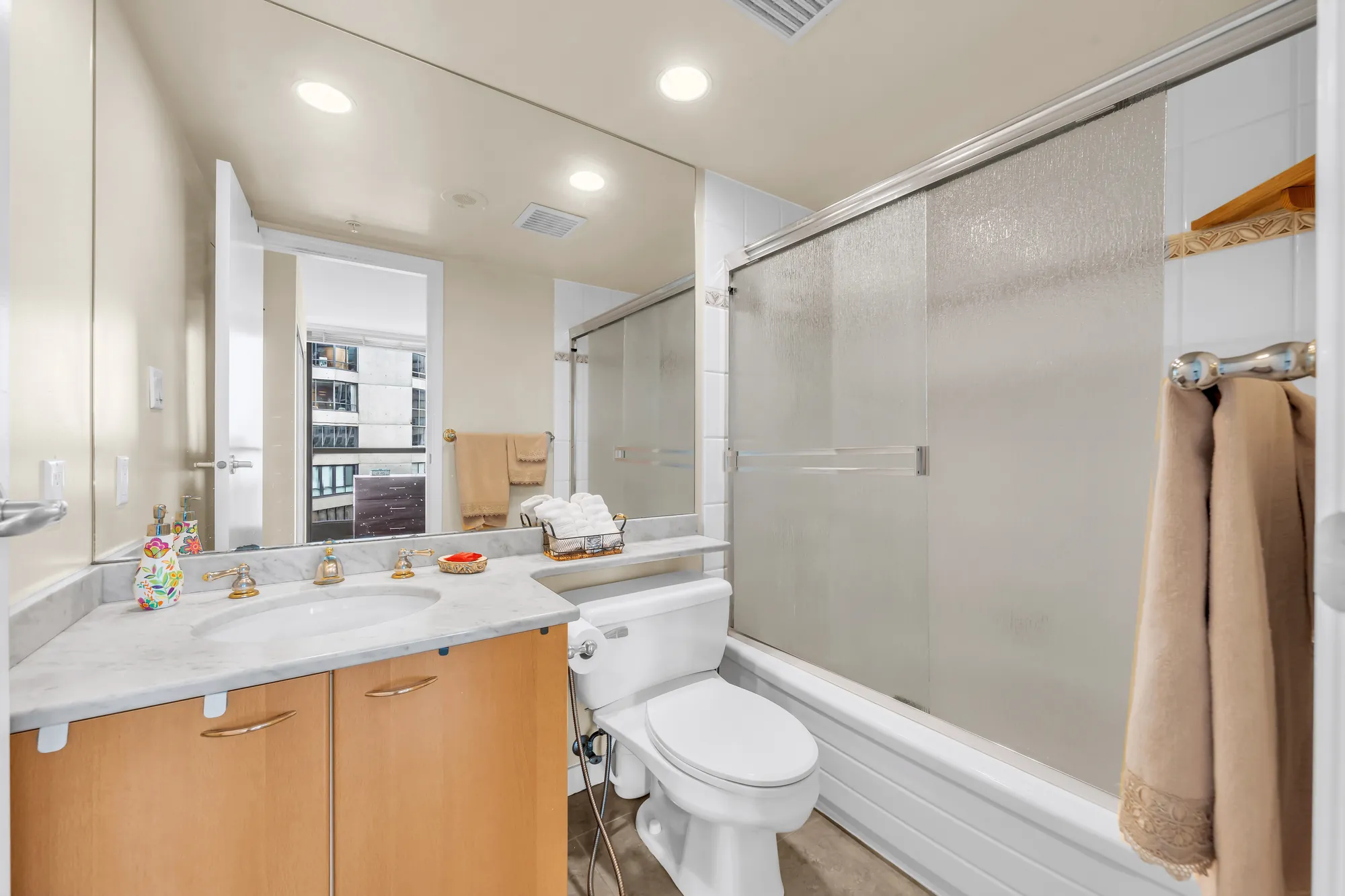 A bathroom: a tub with a frosted sliding glass door, a marble-look counter on a maple vanity, a toilet, a mirror reflecting the door to the bedroom