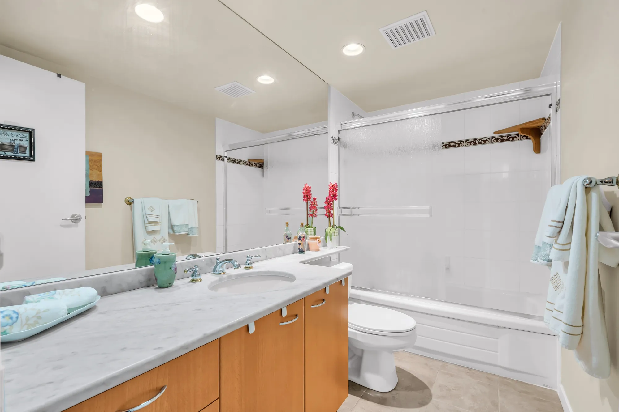 The second bathroom: a long marble-look counter on maple cabinets, a wide mirror, a tub with a sliding glass door, a toilet