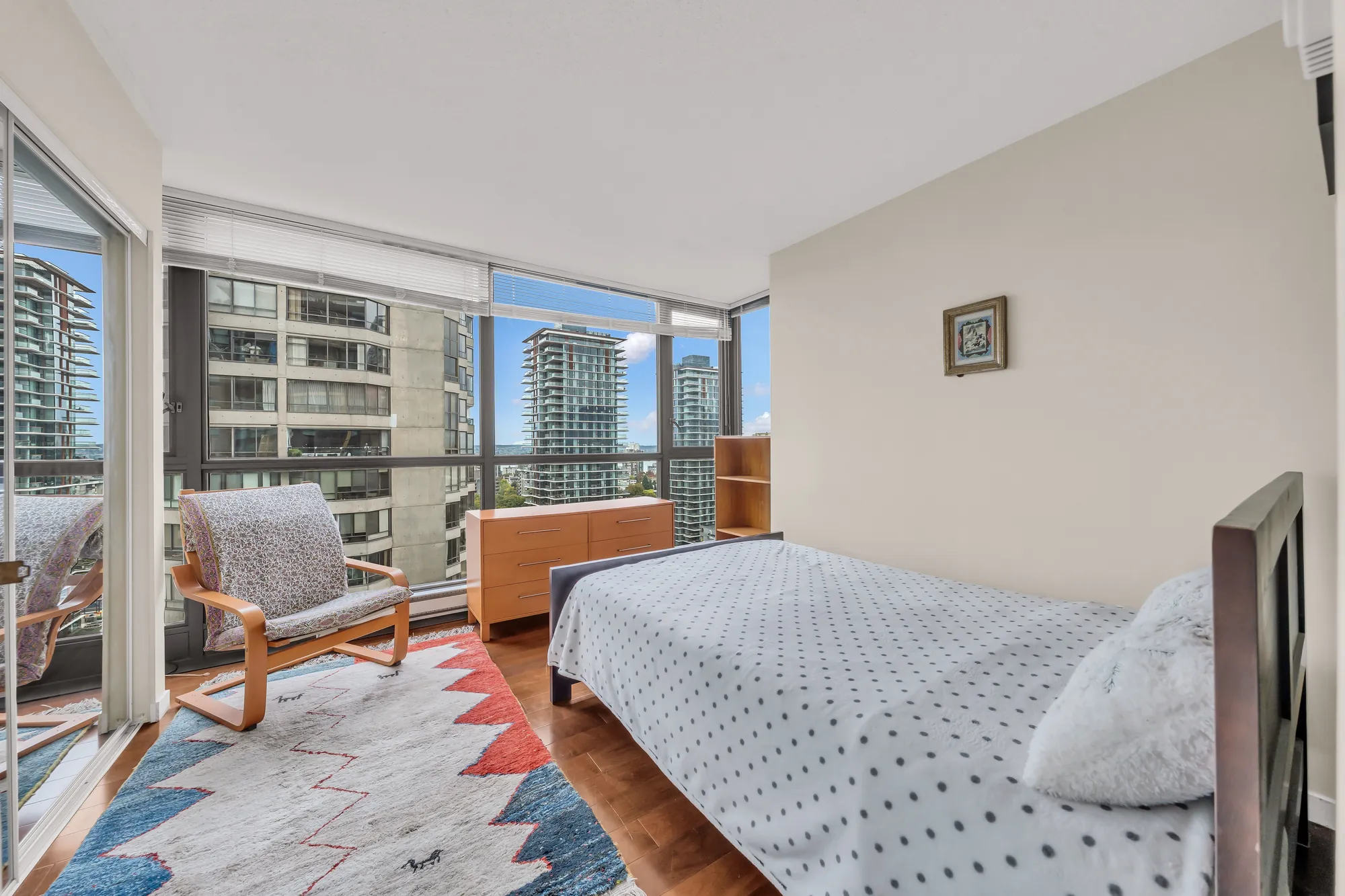 A bedroom with floor-to-ceiling windows on two sides: a bed, a wood armchair, a low cabinet and a patterned rug, the towers beyond