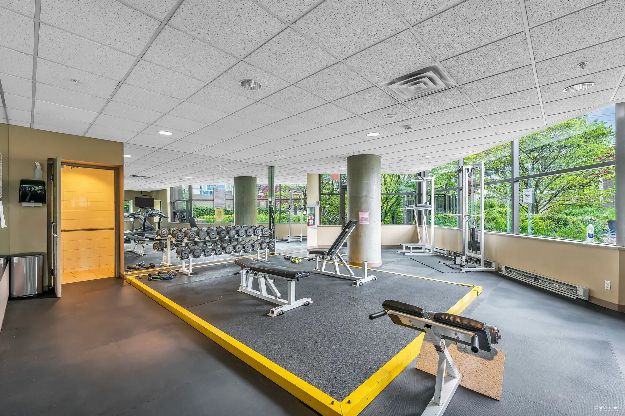 The exercise room from the other end: a bench, free weights, machines, windows along the wall