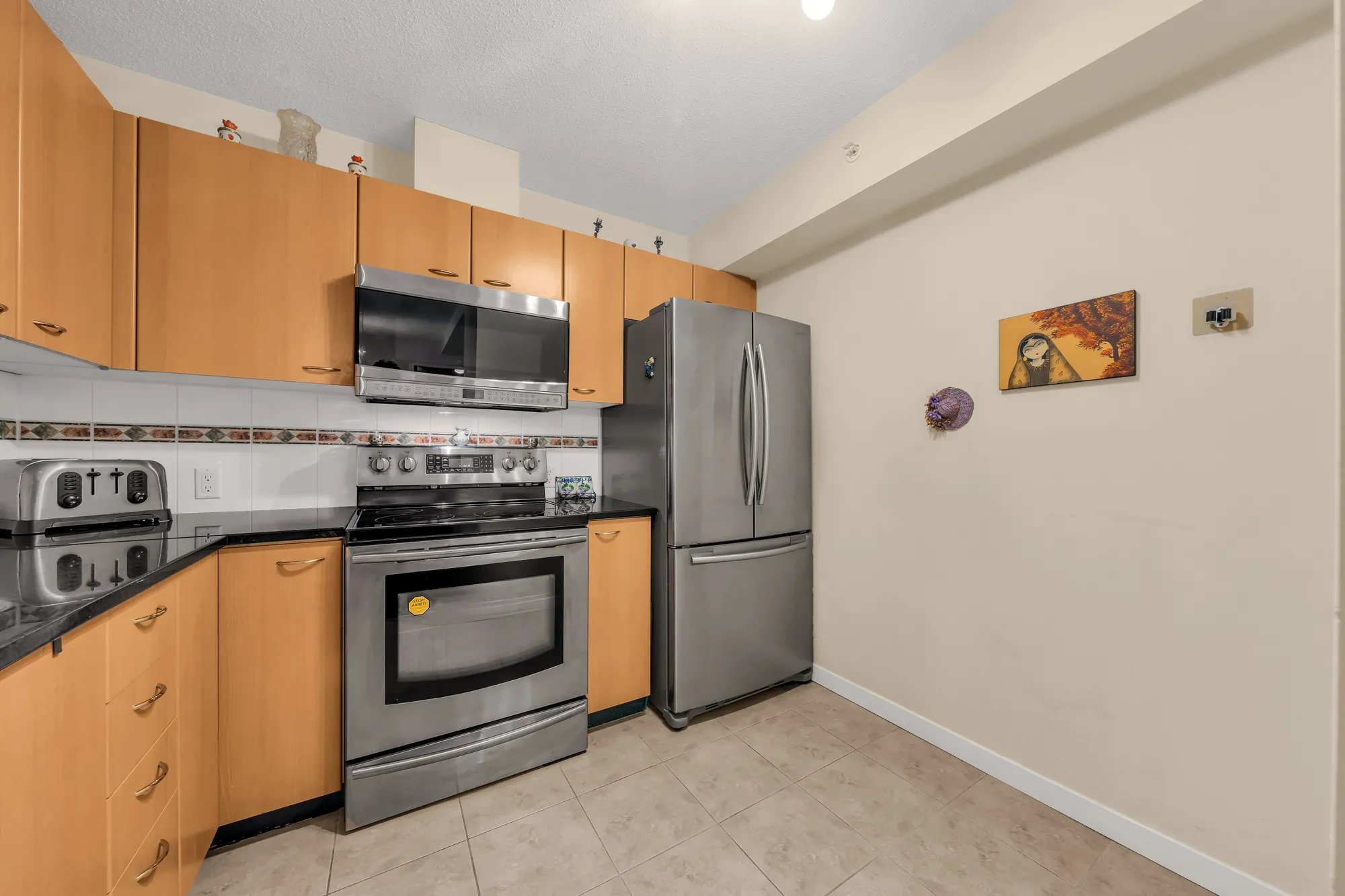 The kitchen: maple cabinets, black counters, a tiled backsplash with a border, a stainless range, microwave and fridge, a tiled floor