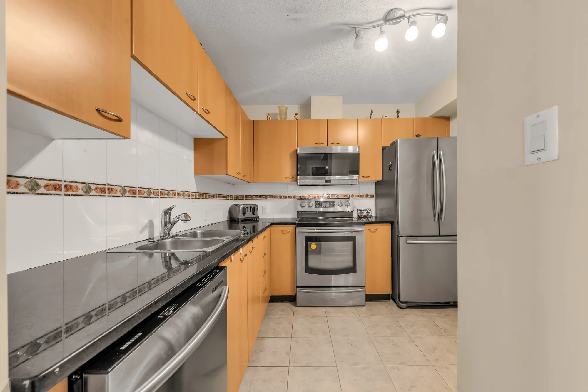 The kitchen counters toward the range and the fridge, pot lights and a track light overhead