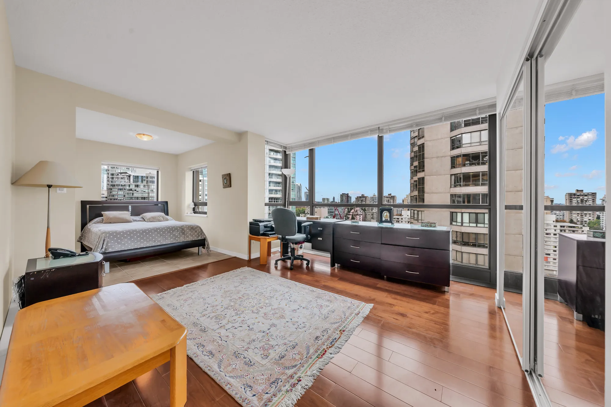 The primary bedroom: a bed with a dark headboard, a rug, a desk area by the floor-to-ceiling windows, hardwood floors