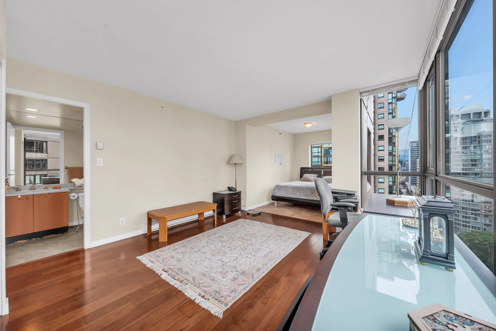 The primary bedroom from the desk end: a glass-topped desk, a bench and a rug, the bed at the far end, windows along the side