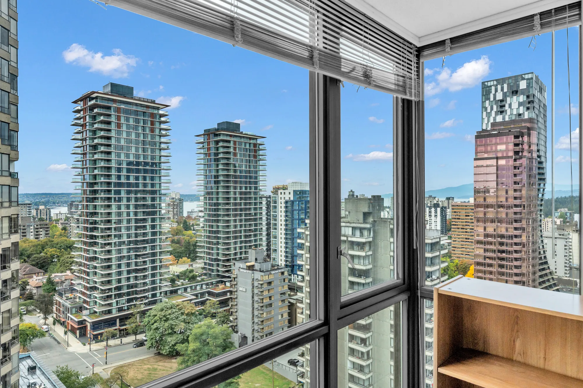 The view from a corner window of 2302-1367 Alberni Street: towers of the West End, the harbour between them and the North Shore mountains under a blue sky